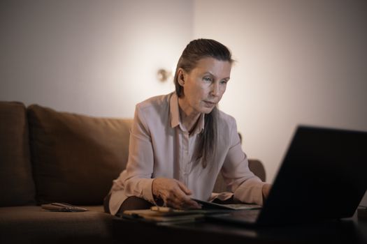 Elderly woman working on laptop in dimly lit living room setting, focused and engaged.