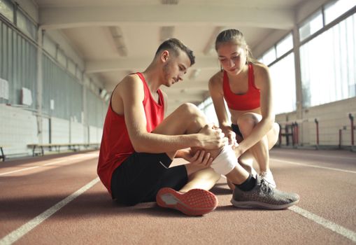 Side view of athlete sitting on rubber track in sports hall while sportive female partner helping to band leg after injury