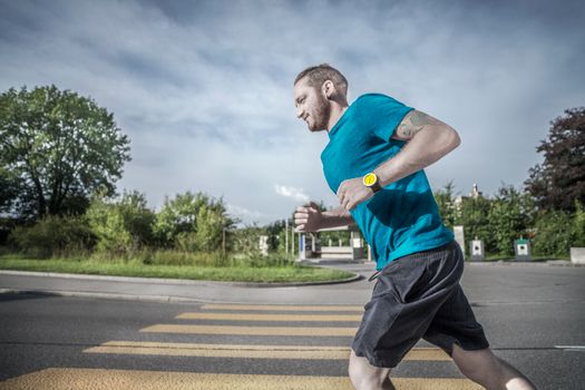 Caucasian man jogging on a city street crosswalk wearing a blue t-shirt and yellow watch.